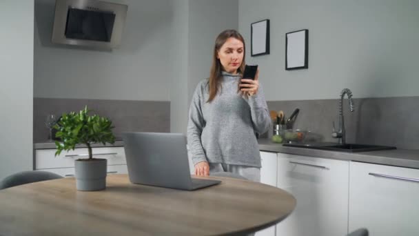 Kobieta ze smartfonem w ręku stojącym w domu przytulne kuchnia używać laptopa do pracy freelance, patrząc skoncentrowany głęboko zagubiony w myślach. Koncepcja pracy zdalnej. Ministerstwo Spraw Wewnętrznych — Wideo stockowe