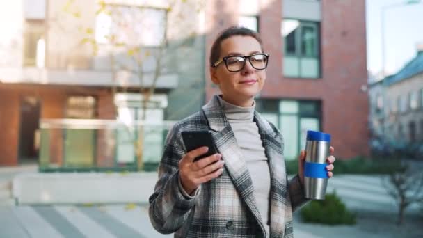 Caucasian businesswoman with glasses and a coat walks through the business district, with thermo cup and using smartphone. — Stock Video