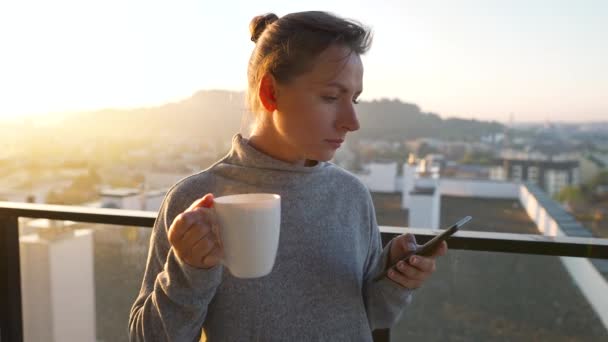 Жінка починає свій день з чашки чаю або кави і перевіряє електронні листи в своєму смартфоні на балконі на світанку, повільний рух . — стокове відео