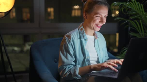 En kvinne sitter i lenestolen og jobber på en bærbar PC om kvelden, eller tekster noen. Fjernarbeidsbegrep. – stockvideo