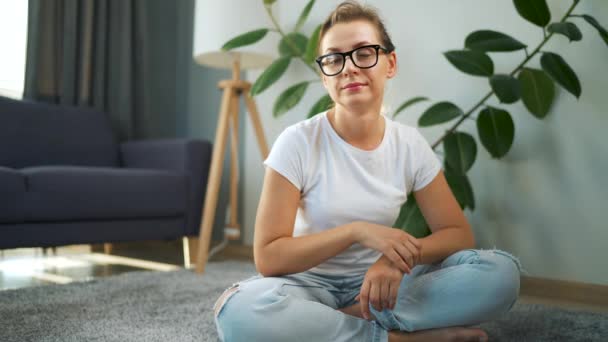 Kvinna med glasögon tittar på kameran sitter på mattan i det inre av en mysig lägenhet. — Stockvideo
