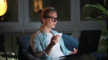 Kadın koltukta oturuyor ve geceleri kredi kartı ve dizüstü bilgisayar kullanarak online alışveriş yapıyor..