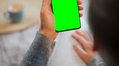 Evde dikey modda yeşil ekranlı akıllı telefon kullanan bir kadın var. Kız internette geziniyor, içeriği izliyor, videolar