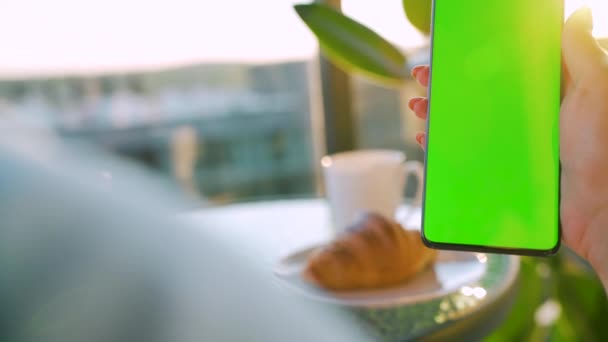 Kvinna sitter i ett café och använder smartphone med grön mock-up skärm i vertikalt läge. Flicka surfa på Internet, titta på innehåll, videor, bloggar. — Stockvideo