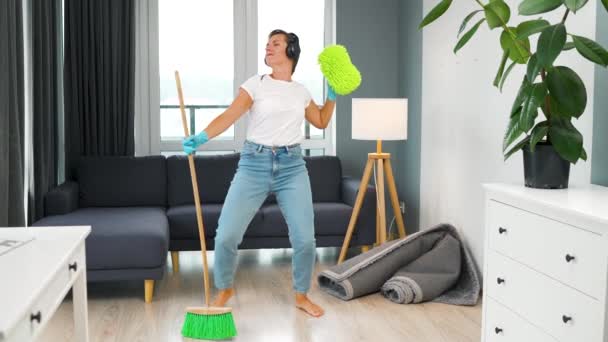 Mujer con auriculares limpiando la casa y divirtiéndose bailando con una escoba y un paño. Movimiento lento — Vídeo de stock