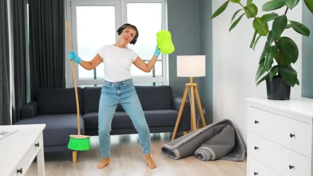 Kobieta w słuchawkach sprzątanie domu i zabawy taniec z miotłą i praniem — Wideo stockowe