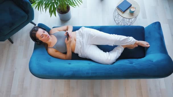 Donna con cuffie wireless che ascolta musica e utilizza app mobili o comunica sui social network su smartphone sdraiata sul divano di casa, vista dall'alto. — Video Stock