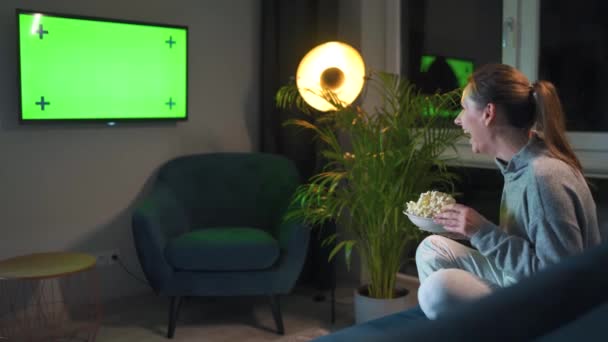 Achteraanzicht van een vrouw zittend op een bank in de woonkamer in de avond en kijkend naar een groene tv-scherm mockup, is blij over wat ze ziet. — Stockvideo