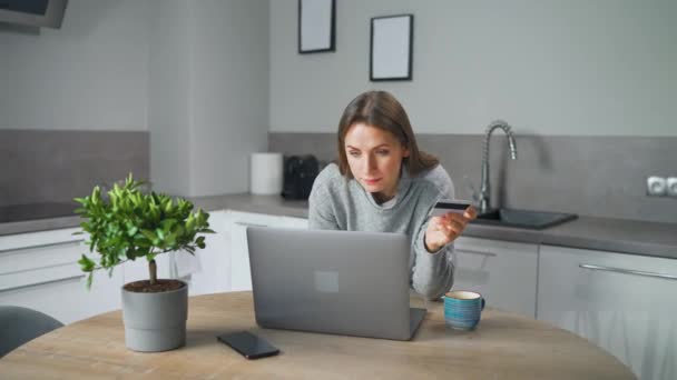 Donna in piedi in casa accogliente cucina, appoggiata sul tavolo e fa shopping online con carta di credito e laptop. — Video Stock