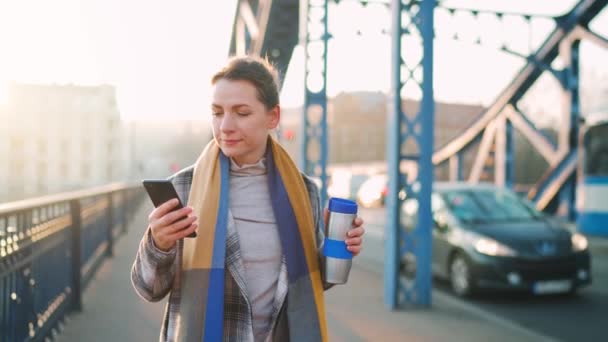 Biała bizneswoman w płaszczu, przechadzająca się przez most w mroźny poranek, pijąca kawę i używająca smartfona. Komunikacja, dzień pracy, pracowite życie. — Wideo stockowe