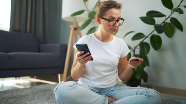 Kobieta w okularach siedzi na podłodze i dokonuje zakupu online za pomocą karty kredytowej i smartfona. Zakupy online, technologia stylu życia — Wideo stockowe