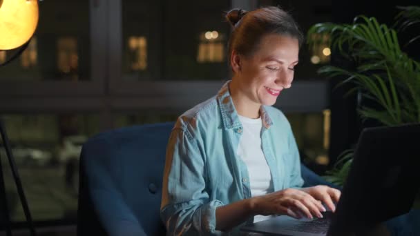 La mujer está sentada en el sillón y trabajando en una computadora portátil por la noche o enviando mensajes de texto a alguien. Concepto de trabajo remoto. — Vídeo de stock