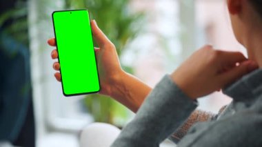 Evde dikey modda yeşil ekranlı akıllı telefon kullanan bir kadın var. Kız internette geziniyor, içeriği izliyor, videolar