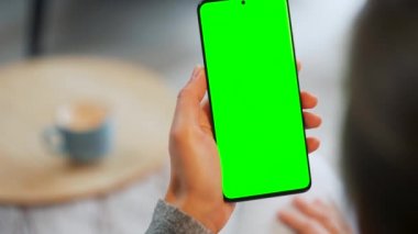 Evde dikey modda yeşil ekranlı akıllı telefon kullanan bir kadın var. Kız internette geziniyor, içeriği izliyor, videolar