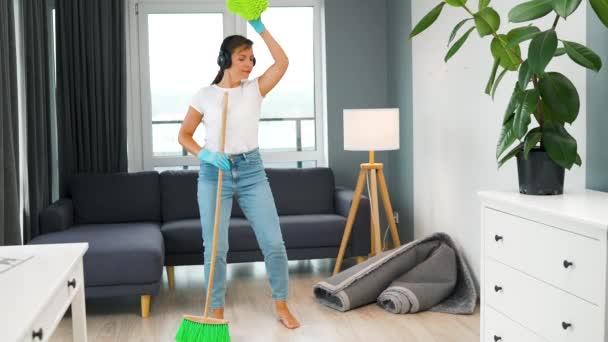 Femme en casque nettoyant la maison et s'amusant à danser avec un balai et un gant de toilette. Mouvement lent — Video