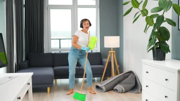ヘッドフォンの女性は家を掃除し、ほうきと洗濯で楽しいダンスをしています。スローモーション — ストック動画