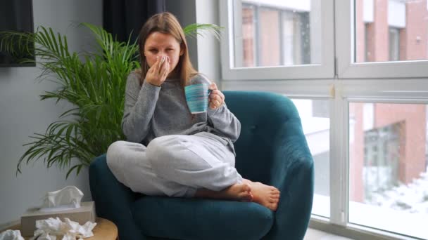 Donna malsana si siede su una sedia, beve tè caldo o una medicina fredda e starnutisce o soffia il naso in un tovagliolo perché ha un raffreddore, influenza, coronavirus. Fuori nevica. — Video Stock