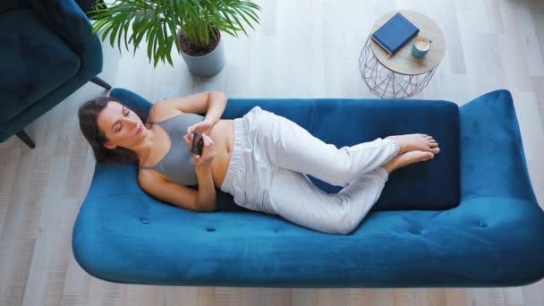 Fotografia aérea de mulher relaxada feliz segurando telefone inteligente, usando aplicativos móveis, assistindo vídeo engraçado, se divertindo conversando nas mídias sociais, deitado no sofá em casa. — Vídeo de Stock