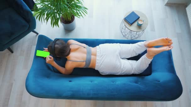 Donna con cuffie wireless che ascolta musica e utilizza app mobili o comunica sui social network su smartphone con schermo modello verde, sdraiata sul divano di casa, vista dall'alto — Video Stock