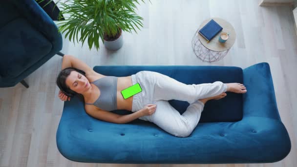Fotografia aérea de mulher relaxada ouvindo palestra ou música em fones de ouvido no smartphone com tela verde mock-up, deitada no sofá em casa. — Vídeo de Stock