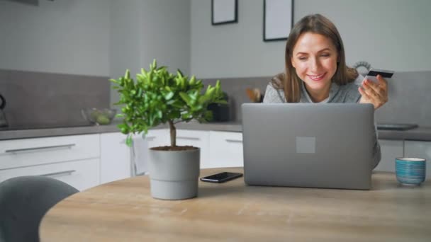 Wanita yang berdiri di dapur yang nyaman di rumah, bersandar di atas meja dan membuat belanja online menggunakan kartu kredit dan laptop. — Stok Video