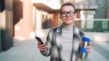 Beyaz tenli, gözlüklü ve ceketli bir iş kadını iş bölgesinde termik bardakla yürüyor ve akıllı telefon kullanıyor..