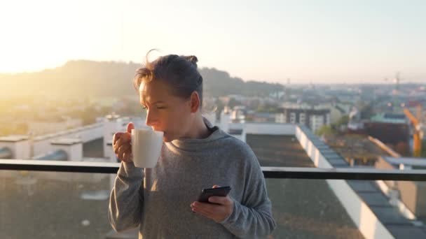 Femme commence sa journée avec une tasse de thé ou de café et vérifier les e-mails dans son smartphone sur le balcon à l'aube. — Video