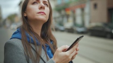 Kadın toplu taşıma durağında duruyor ve zaman çizelgesini kontrol ediyor. Tramvay arka planda duruyor..