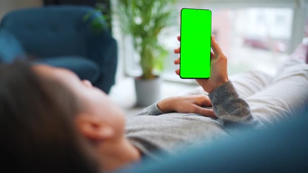 Vrouw ligt thuis op de bank en gebruikt smartphone met groene mock-up scherm in verticale modus. Meisje surfen op internet, kijken naar inhoud, video 's, blogs. — Stockvideo