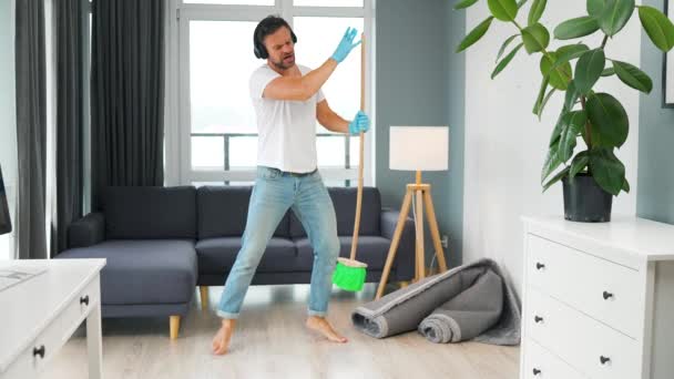 Man in headphones cleaning the house and having fun dancing with a broom. Slow motion — Vídeo de stock
