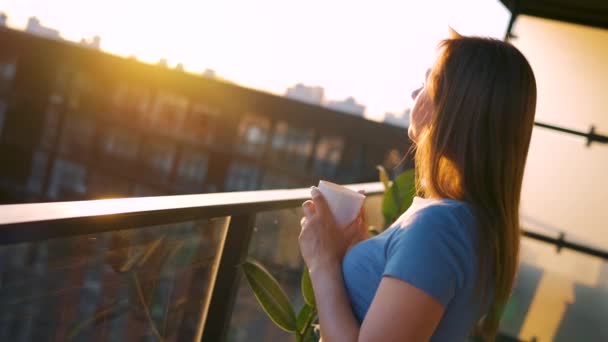 Frau trinkt Kaffee oder Tee, während sie auf dem Balkon steht und den Sonnenuntergang bewundert. — Stockvideo