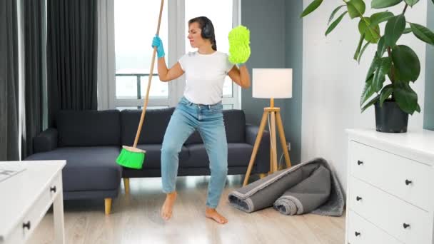 Γυναίκα με ακουστικά που καθαρίζει το σπίτι και διασκεδάζει χορεύοντας με σκούπα και πετσέτα. Αργή κίνηση — Αρχείο Βίντεο