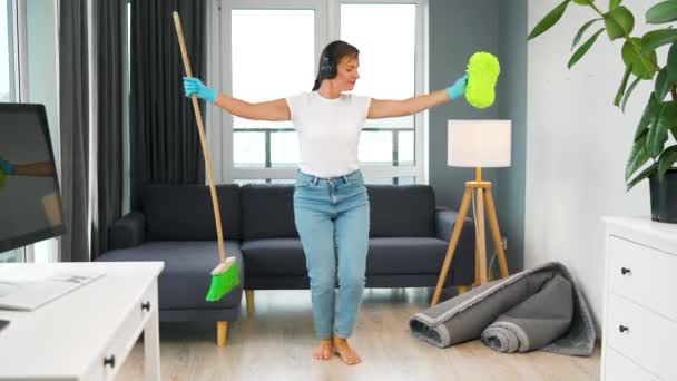 ヘッドフォンの女性は家を掃除し、ほうきと洗濯布で楽しいダンスをしています. — ストック動画