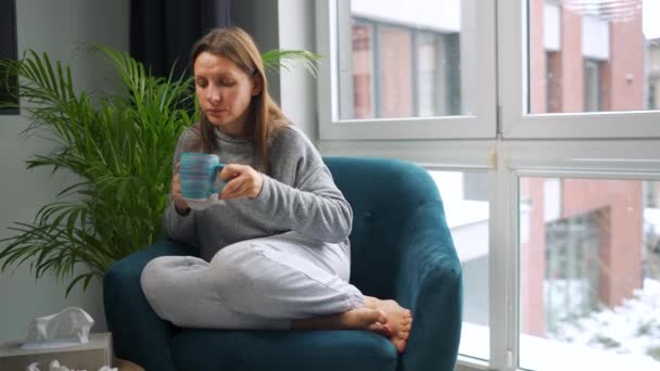 Niezdrowa kobieta siedzi na krześle, pije gorącą herbatę lub lek na przeziębienie i kicha lub wbija nos w serwetkę, bo ma przeziębienie, grypę, koronawirus. Na zewnątrz pada śnieg. — Wideo stockowe