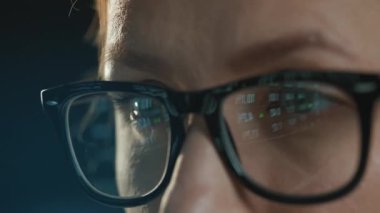 Gözlüklü kadın monitöre bakıyor ve veri ve analizlerle çalışıyor. Monitör ekranı gözlüklere yansıyor. Gece çalış. Aşırı yakın çekim