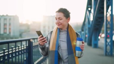 Beyaz ceketli bir iş kadını, soğuk bir sabahta köprüde yürüyor, kahve içiyor ve akıllı telefon kullanıyor. İletişim, iş günü, yoğun yaşam konsepti.