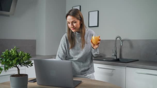 Woman with a glass of juice in her hand stands in the kitchen and works at a laptop do freelance work, chatting with someone, answering e-mail. Concept of remote work. — Stock video