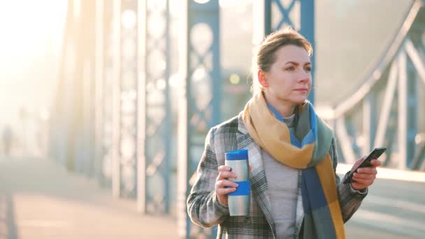 Retrato de una joven mujer de negocios caucásica en un abrigo, caminando por la ciudad en una mañana helada, tomando café y usando un teléfono inteligente. Comunicación, día de trabajo, concepto de vida ocupada. Movimiento lento — Vídeo de stock