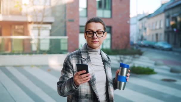 Caucasian businesswoman with glasses and a coat walks through the business district, with thermo cup and using smartphone. Communication, work day, busy life concept — Stock Video
