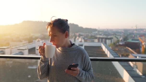 Femme commence sa journée avec une tasse de thé ou de café et vérifier les e-mails dans son smartphone sur le balcon à l'aube, au ralenti. — Video