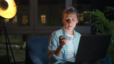 Kadın koltukta oturuyor ve geceleri kredi kartı ve dizüstü bilgisayar kullanarak online alışveriş yapıyor..