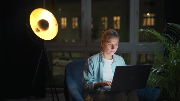 Kobieta siedzi w fotelu i pracuje w nocy nad laptopem albo pisze do kogoś. Pojęcie pracy zdalnej. — Wideo stockowe