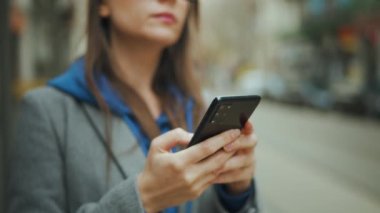 Woman with glasses stands at a public transport stop and uses smartphone. Tram passes in the background.