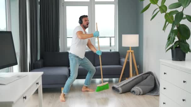 Man in headphones cleaning the house and having fun dancing with a broom. Slow motion — Wideo stockowe