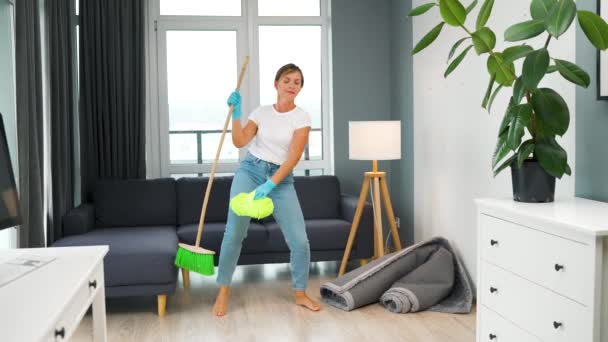 ヘッドフォンの女性は家を掃除し、ほうきと洗濯で楽しいダンスをしています。スローモーション — ストック動画