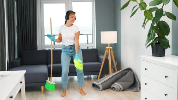 Γυναίκα με ακουστικά που καθαρίζει το σπίτι και διασκεδάζει χορεύοντας με σκούπα και πετσέτα. Αργή κίνηση — Αρχείο Βίντεο