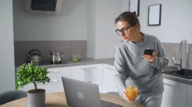 Elinde meyve suyu olan bir kadın mutfakta duruyor ve dizüstü bilgisayarda çalışıyor. Serbest çalışıyor, biriyle sohbet ediyor, e-postalara cevap veriyor. Uzak çalışma kavramı.