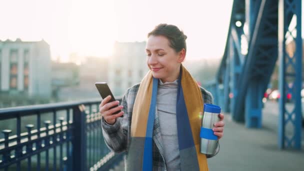 Biała bizneswoman w płaszczu, przechadzająca się przez most w mroźny poranek, pijąca kawę i używająca smartfona. Komunikacja, dzień pracy, pracowite życie. — Wideo stockowe