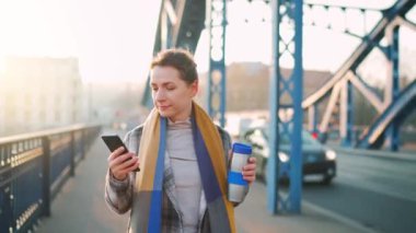 Beyaz ceketli bir iş kadını, soğuk bir sabahta köprüde yürüyor, kahve içiyor ve akıllı telefon kullanıyor. İletişim, iş günü, yoğun yaşam konsepti.