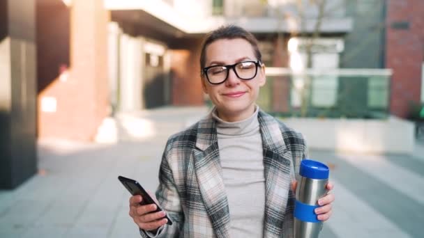 Mujer de negocios caucásica con gafas y un abrigo camina por el distrito de negocios, con termo taza y el uso de teléfono inteligente. Comunicación, día de trabajo, concepto de vida ocupada — Vídeos de Stock